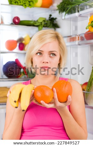 woman looking sad hold banana orange in hands negative emotion near open door refrigerator, concept of diet health food, pretty girl hold vitamin fruit