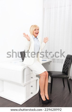 Business woman excited hold hands up raised arms sitting at modern office desk, surprised happy smile businesswoman success