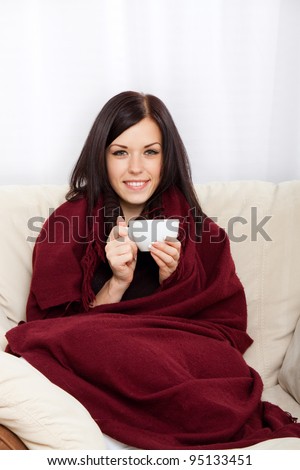 Portrait of smiling beautiful pretty young woman drinking hot coffee at home - indoors covered with blanket holding cup of tea, sitting on sofa, couch