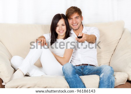 Couple watching tv in their living room at home, young happy smile man and woman sitting on couch, sofa, hold remote control