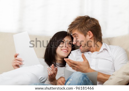 Happy couple reading a letter in their living room, young smile man and woman sitting on couch, sofa looking to each other talking