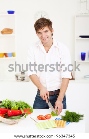 portrait of handsome young man slice, cutting green pepper, tomato in the kitchen, cooking, prepare  vegetable salad, happy smile, looking at camera