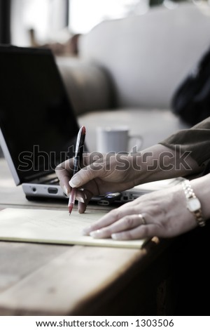 Businesswoman Writing Notes next to her Laptop Computer in a Hotel Atrium (slight color, shallow focus point on hand with pen).