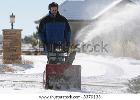 Man using a powerful snow blower in wintertime, with a large house in the background (shallow focus point on the snow blower).