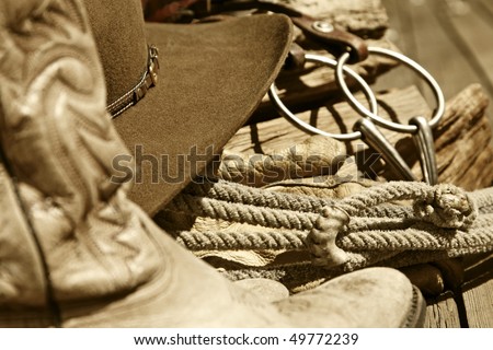 Sepia-toned rustic western image of cowboy boots, cowboy hat, rope, horse bit and stacked wood (mid-focus point).