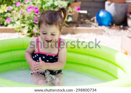 A little toddler girl in a black swimsuit playing in a green paddling pool in the garden