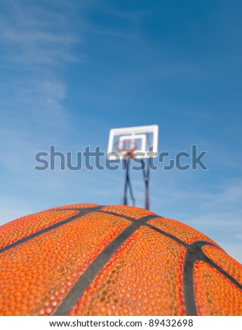 Basketball and a street basket back board with hoop in the background against blue sky. Shallow depth of field, focusing on ball\'s worn surface. Copy space for your text.