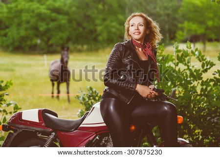 Biker girl in leather jacket on a motorcycle over the background of horse. Warm toned image