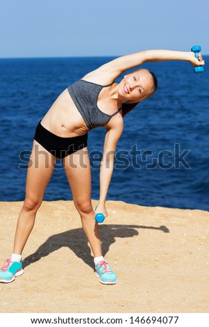 Portrait of healthy young woman in sportswear exercising with hand weights on the beach