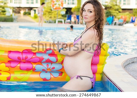 Beautiful pregnant woman with colorful mattress near swimming pool with blue water, summer time