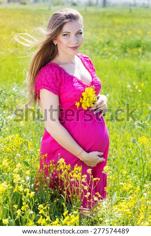 Happy pregnant woman is wearing pink dress on a field with yellow flowers, pregnancy girl