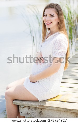 Happy pregnant woman is wearing white dress sitting on bridge near blue lake, summer time, pregnancy girl