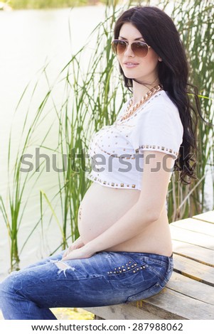 Happy pregnant girl is wearing jeans and white shirt is sitting on wooden bridge near blue river, summer time, pregnancy girl