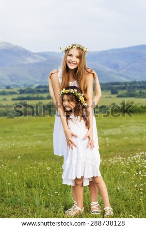 Cute sisters girls are wearing white dresses at  green camomile field with mountains view