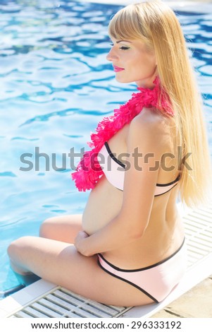 Beautiful pregnant woman is wearing pink hawaiian flowers sitting near swimming pool with blue water