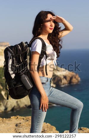 Pretty slim traveler girl is looking to the sea on rock edge over mountains view with backpack, summer time