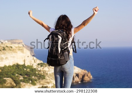 Back view of young pretty slim woman standing with backpack on cliff\'s edge and looking into sea