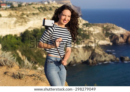 Tourist smiling girl making selfie photo with stick on the peak of mountain over sea background