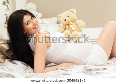 Beautiful pregnant woman is lying on bed and hugging toy teddy bear