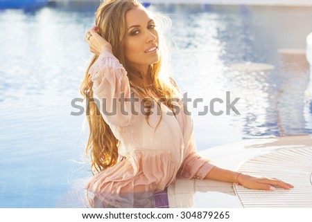 Sexy smiling woman with tanned skin and long shiny hair is wearing wet shirt swimming in pool, summer time
