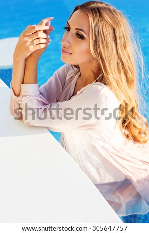Sexy pretty woman with tanned skin and long shiny hair is wearing wet shirt sitting in swimming pool, summer time