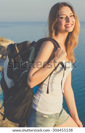 Pretty happy traveler girl is standing on rock edge over sea view with backpack, summer time