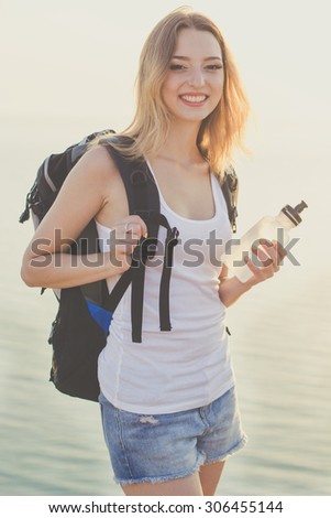 Pretty happy traveler girl is standing on rock edge with water bottle over sea view with backpack, summer time