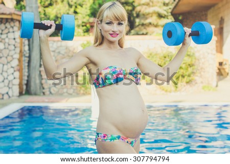 Beautiful pregnant woman is doing sports with dumbbells near swimming pool with blue water