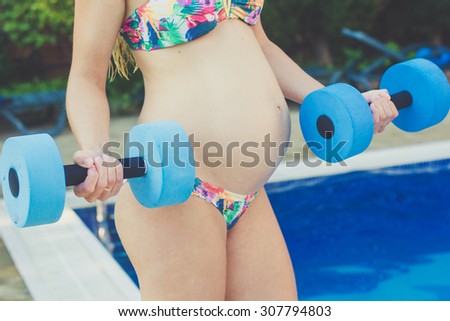 Beautiful pregnant woman is water aerobics with blue lightweight dumbbells near swimming pool