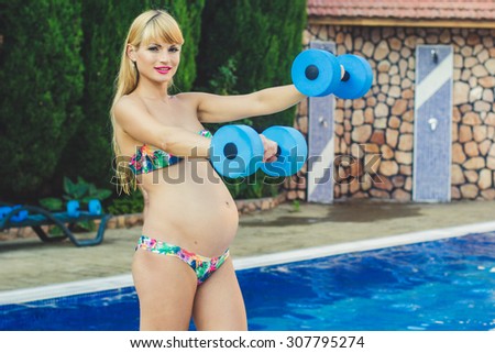Beautiful pregnant woman is doing sports exarcises with dumbbells near swimming pool with blue water