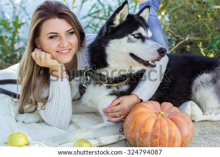 Young pretty girl and her blueyed dog husky is lying outdoors near lake with pumpkin and apples, autumn time