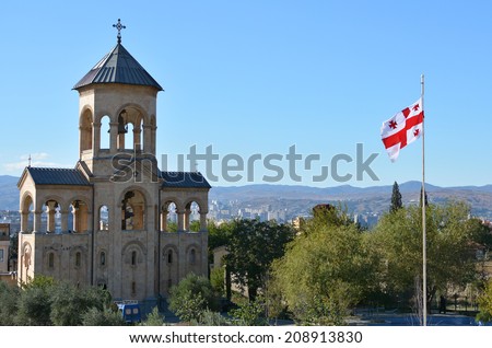 Tbilisi, Georgia - October 7, 2013: Georgian flag fluttering in the wind on the territory of the Holy Trinity Cathedral (Tsminda Sameba), Caucasus Range and panorama of Tbilisi
