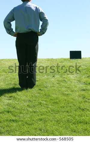 Young man, with hands on hips, looks at laptop that is ten feet away, against a blue, cloudless sky and on green, grassy lawn