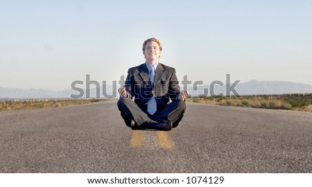 Business man sitting in the middle of the road, facing the camera meditating, in a black suit, in the middle of the desert.