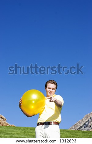 Casual white man points finger at the screen with a small smile on his face with a yellow ball in his hands under a blue sky