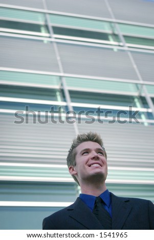 Blond hair blue eye business man is smiling wearing a black suit, blue shirt, and balck tie against a glass office building