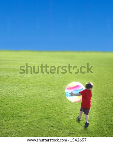 Young boy holding large ball as he walks on the green grass against the blue sky