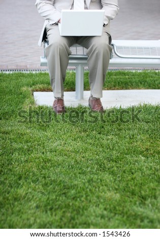 Businessman is sitting on metal bench next to the grass in his tan suit as he types on his laptop