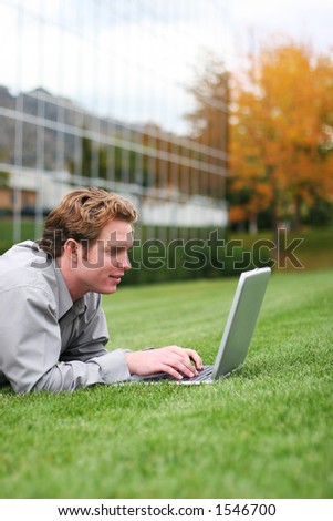 Businessman with gray shirt lies down in green grass as he types on his laptop with glass office building behind him