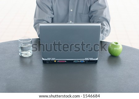 Businessman in gray shirt types on his laptop with green apple and glass of water on his sides