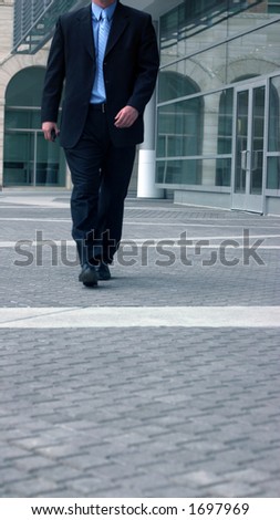 Business man in darker suit with blue shirt and blue tie walks forward next to office building