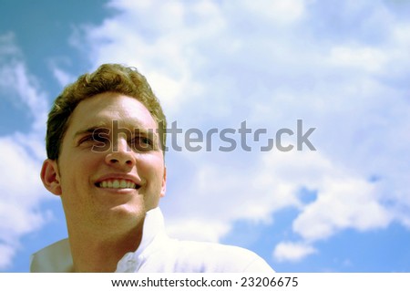 A young, happy man looks over his shoulder while wearing a white shirt with and with a big smile while standing under a blue sky full of clouds