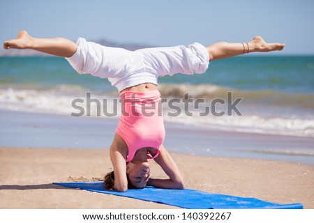 Beautiful young woman standing on her head with her legs spread as part of a yoga pose at the beach