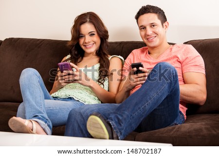 Happy couple updating their social network statuses on their cell phone while relaxing in the living room