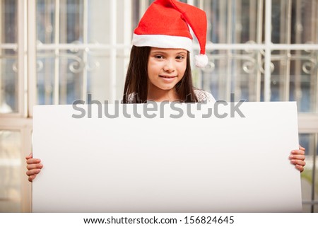 Happy little girl wearing Santa\'s hat and holding a big sign at home