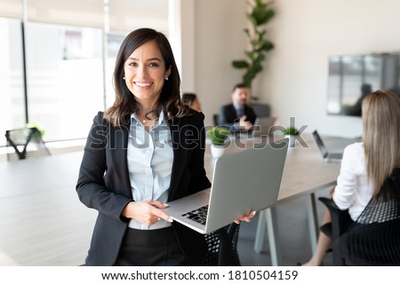 Portrait of smiling female entrepreneur holding a laptop with team in background at office conference room