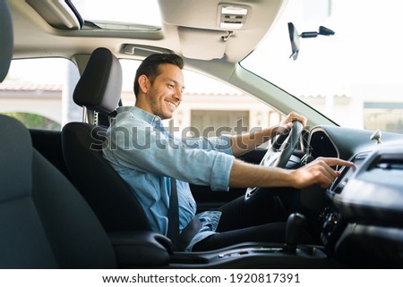 Handsome man in his 30s sitting in the driver's seat and smiling. Taxi driver listening to music on the car and changing the radio station