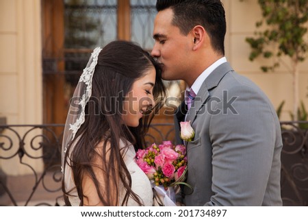 Handsome Latin groom kissing her beautiful bride in the forehead outside a church