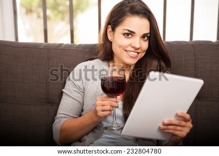 Cute Latin girl having a glass of wine at home and reading on a tablet computer