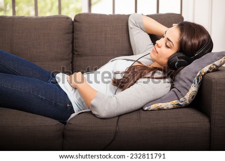 Pretty young brunette laying on her couch and listening to music using headphones